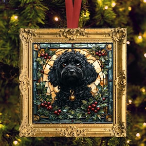 Puede incluir: Adorno navideño cuadrado con marco dorado que presenta un perro negro con collar dorado. El perro está rodeado de acebo y bayas rojas, sobre un fondo de vidrieras. Se adjunta una cinta roja para colgar.