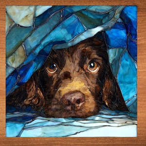 May include: A brown dog peeks out from under a blue and white stained glass-like design. The dog has brown fur, a tan muzzle, and large, expressive brown eyes. The artwork is set against a wooden background.