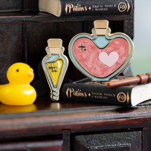 May include: Three gold-toned enamel pins depicting potion bottles. The heart-shaped bottle is filled with pink glitter and has a white heart inside. The other two bottles are filled with yellow and green glitter. The pins are sitting on a black book with the title "Potions" and a yellow rubber duck is to the left of the pins.