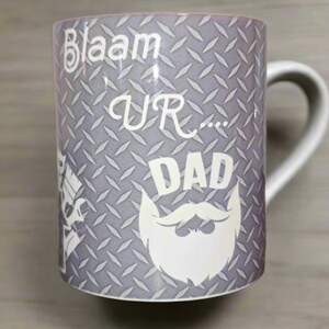 May include: A gray ceramic mug with a white handle. The mug features a diamond plate pattern and the words "Blaam UR... DAD" in white, along with a white beard graphic. The mug is designed for Father's Day.