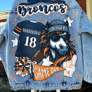 Könnte beinhalten: Hellblaue Jeansjacke mit individuellem Design. Auf der Rückseite der Jacke befindet sich das Wort "Broncos" in Marineblau und Orange, ein Football-Trikot mit "Manning 18", ein Pferd mit Sonnenbrille und Mütze sowie ein "Game Day" Banner.