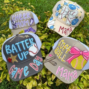 May include: Four baseball and softball themed trucker caps with mesh backs. The caps are tan, grey, and white, with hand-painted designs and text including "Cheer Mom", "Mama", "Batter Up", "Game Day", and "Softball Mama".