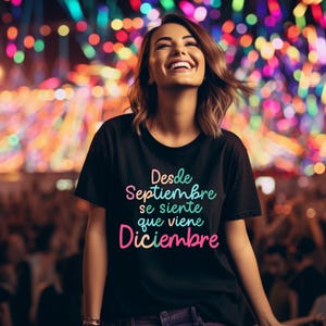 May include: A black t-shirt with the text "Desde Septiembre se siente que viene Diciembre" in colorful gradient lettering. The person wearing the shirt is smiling and looking upwards, with a blurred background of colorful lights.