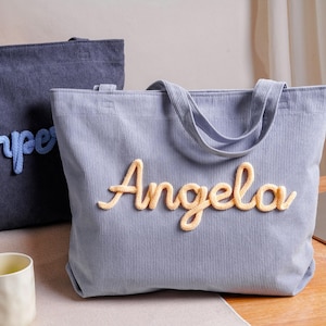 May include: A light blue corduroy tote bag featuring the name Angela in raised, yellow, yarn-like script lettering. A second, darker blue corduroy tote bag with the name Harper in light blue script lettering is positioned behind the first bag.