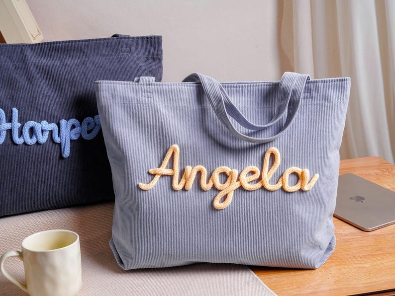 May include: A light blue corduroy tote bag featuring the name Angela in raised, yellow, yarn-like script lettering. A second, darker blue corduroy tote bag with the name Harper in light blue script lettering is positioned behind the first bag.