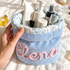 May include: A light blue corduroy storage basket featuring the name Elena embroidered in pink yarn on the front. The interior is lined with a white fabric patterned with small blue flowers and contains various skincare bottles and tubes.