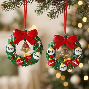K&ouml;nnte beinhalten: Zwei runde Weihnachtskranz-Anh&auml;nger mit gr&uuml;nem Blattwerk, roten Schleifen und goldenen Glocken. Jeder Kranz ist mit kleinen rot-wei&szlig;en Zuckerstangen, roten Beeren sowie wei&szlig;en oder roten Kugeln mit Namen und der Jahreszahl 2026 verziert.