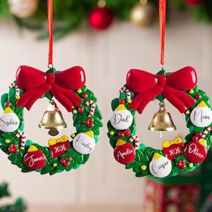 K&ouml;nnte beinhalten: Zwei kreisf&ouml;rmige Weihnachtsbaumschmuck-Kr&auml;nze mit gr&uuml;nen Stechpalmenbl&auml;ttern, einer gro&szlig;en roten Schleife oben und einer goldenen Glocke in der Mitte. Jeder Kranz ist mit kleinen roten und wei&szlig;en Kugeln verziert, die mit den Namen Sophia, James, Kinsley, Dad, Mom, Amelia, Cooper, Oliver und der Jahreszahl 2025 beschriftet sind.