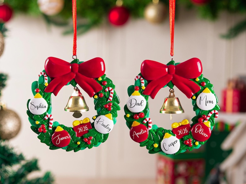 K&ouml;nnte beinhalten: Zwei kreisf&ouml;rmige Weihnachtsbaumschmuck-Kr&auml;nze mit gr&uuml;nen Stechpalmenbl&auml;ttern, einer gro&szlig;en roten Schleife oben und einer goldenen Glocke in der Mitte. Jeder Kranz ist mit kleinen roten und wei&szlig;en Kugeln verziert, die mit den Namen Sophia, James, Kinsley, Dad, Mom, Amelia, Cooper, Oliver und der Jahreszahl 2025 beschriftet sind.