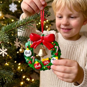K&ouml;nnte beinhalten: Ein runder Weihnachtsbaumschmuck in Form eines Kranzes mit gr&uuml;nem Blattwerk, einer gro&szlig;en roten Schleife oben und einer goldenen Glocke in der Mitte. Der Kranz ist mit roten Beeren, Zuckerstangen und kleinen Geschenken verziert. Er tr&auml;gt die Aufschriften Dad, Noah und 2026.
