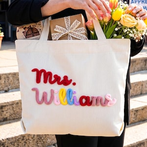 May include: A cream-colored corduroy tote bag featuring the text Mrs. Williams embroidered in raised, multi-colored yarn. The word Mrs. is in red, while the word Williams is spelled out in alternating pink, yellow, blue, purple, and red yarn.