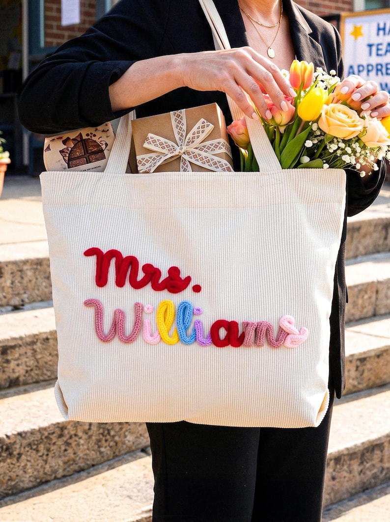 May include: A cream-colored corduroy tote bag featuring the text Mrs. Williams embroidered in raised, multi-colored yarn. The word Mrs. is in red, while the word Williams is spelled out in alternating pink, yellow, blue, purple, and red yarn.