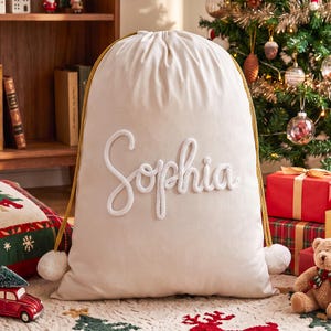 May include: A cream-colored fabric drawstring gift bag featuring the name Sophia in white rope-style lettering across the center. The bag includes gold-colored braided drawstrings with white pom-pom ends, creating a festive storage container.