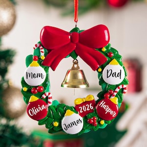 K&ouml;nnte beinhalten: Ein gr&uuml;ner Weihnachtskranz-Anh&auml;nger mit einer gro&szlig;en roten Schleife oben und einer goldenen Glocke in der Mitte. Der Kranz ist mit rot-wei&szlig;en Kugeln mit den Namen Mom, Dad, Olivia, James und Harper sowie einem Geschenk mit der Aufschrift 2026 verziert.