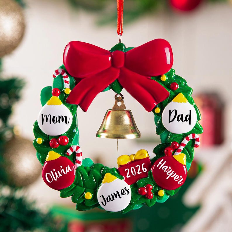 K&ouml;nnte beinhalten: Ein gr&uuml;ner Weihnachtskranz-Anh&auml;nger mit einer gro&szlig;en roten Schleife oben und einer goldenen Glocke in der Mitte. Der Kranz ist mit rot-wei&szlig;en Kugeln mit den Namen Mom, Dad, Olivia, James und Harper sowie einem Geschenk mit der Aufschrift 2026 verziert.