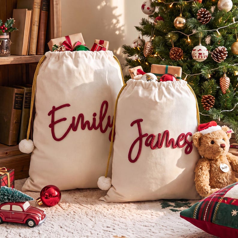 May include: Two off-white canvas drawstring gift bags with gold cords and white pom-poms. The left bag features the name Emily in red script, and the right bag features the name James in red script. Both bags are filled with wrapped gifts.