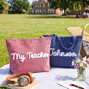May include: Two corduroy tote bags, one dusty pink and one navy blue, each featuring white embroidered script text. The pink bag displays the words My Teacher, while the navy blue bag displays the name Johnson. Both bags have two shoulder straps.
