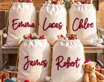 May include: Five cream-colored fabric drawstring sacks with gold cords and white pom-poms, each featuring a name embroidered in red script: Emma, Lucas, Chloe, James, and Robert. The sacks are filled with wrapped gifts and holiday ornaments.