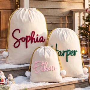 May include: Three off-white drawstring gift bags with gold cords and white pom-poms. The bags feature personalized names in raised, textured script: Sophia in dark red, Harper in dark green, and Ethan in light pink, all in cursive.