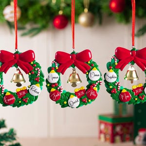 K&ouml;nnte beinhalten: Drei kreisf&ouml;rmige Weihnachtsbaumschmuck-Kr&auml;nze, die an roten B&auml;ndern h&auml;ngen. Jeder gr&uuml;ne Kranz hat eine gro&szlig;e rote Schleife oben, eine goldene Glocke in der Mitte und kleine dekorative Kugeln mit Namen wie Noah, Emma, Robert, Mama, Nana, Jack, David, Alice, Sophia, Max, Bella und Asher sowie die Jahreszahl 2025.