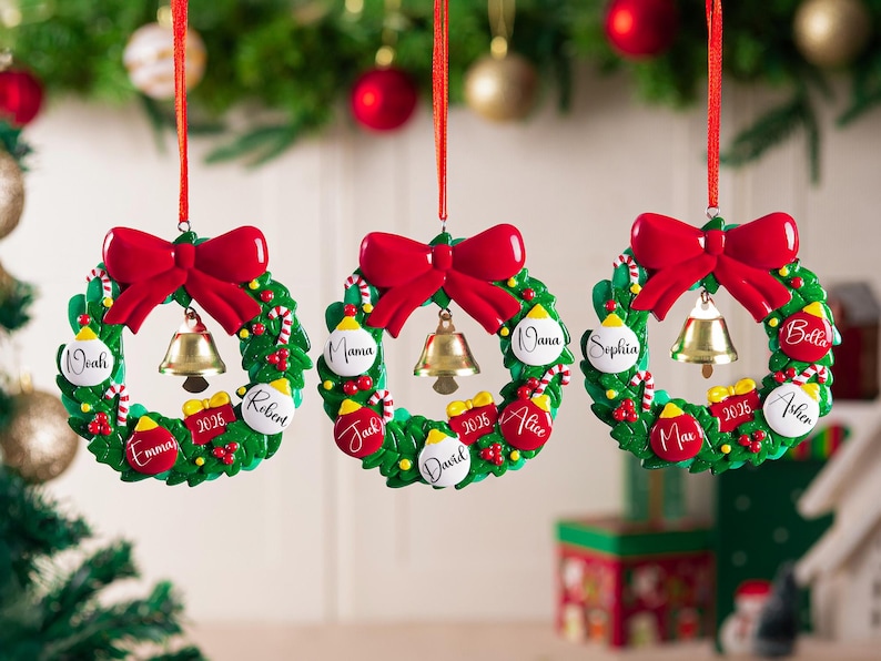 K&ouml;nnte beinhalten: Drei kreisf&ouml;rmige Weihnachtsbaumschmuck-Kr&auml;nze, die an roten B&auml;ndern h&auml;ngen. Jeder gr&uuml;ne Kranz hat eine gro&szlig;e rote Schleife oben, eine goldene Glocke in der Mitte und kleine dekorative Kugeln mit Namen wie Noah, Emma, Robert, Mama, Nana, Jack, David, Alice, Sophia, Max, Bella und Asher sowie die Jahreszahl 2025.