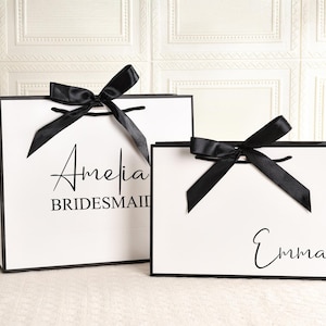 May include: Two white gift bags with black trim and black satin bows. One bag has the name "Amelia" and the word "Bridesmaid" printed on it. The other bag has the name "Emma" printed on it. The bags are on a white surface.