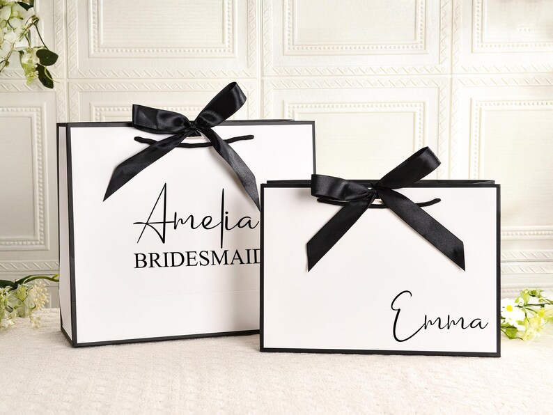 May include: Two white gift bags with black trim and black satin bows. One bag has the name "Amelia" and the word "Bridesmaid" printed on it. The other bag has the name "Emma" printed on it. The bags are on a white surface.