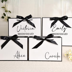 May include: White gift bags with black trim and black satin bows. Each bag has a name printed in elegant script: Victoria, Emily, Alice, and Camila. The word "Bridesmaid" is printed below Victoria and Emily's names.