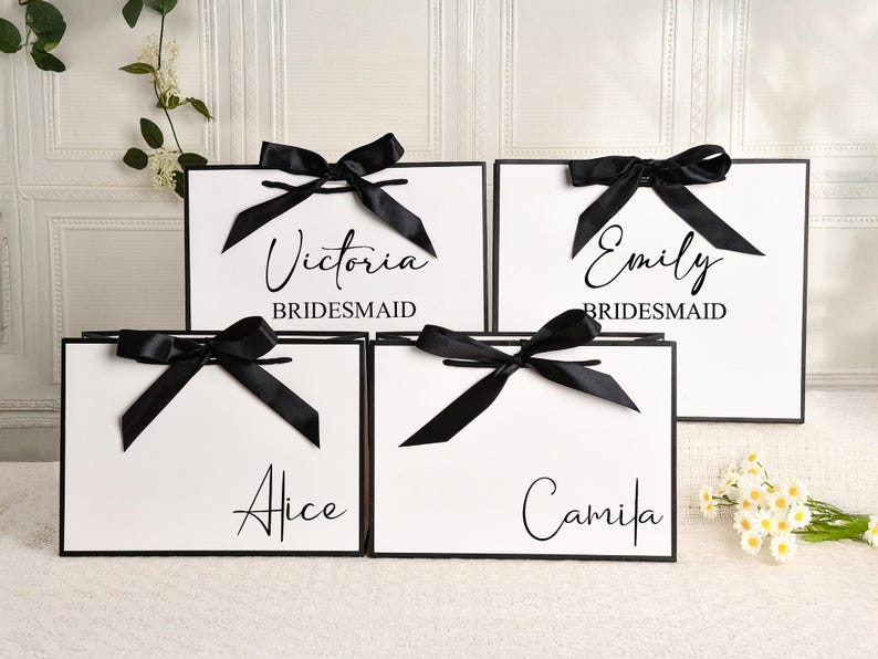 May include: White gift bags with black trim and black satin bows. Each bag has a name printed in elegant script: Victoria, Emily, Alice, and Camila. The word "Bridesmaid" is printed below Victoria and Emily's names.