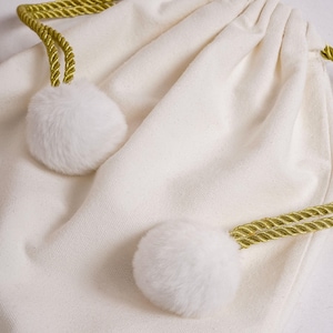 May include: An off-white drawstring pouch made of canvas fabric. The bag features two gold-colored twisted rope drawstrings, each ending in a soft, fluffy white faux fur pom-pom. The pouch is gathered at the top by the drawstrings.