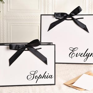 May include: Two white gift bags with black trim and black satin bows. One bag has the name "Sophia" in cursive, and the other has "Evelyn". The bags are on a white surface with a floral and card in the background.