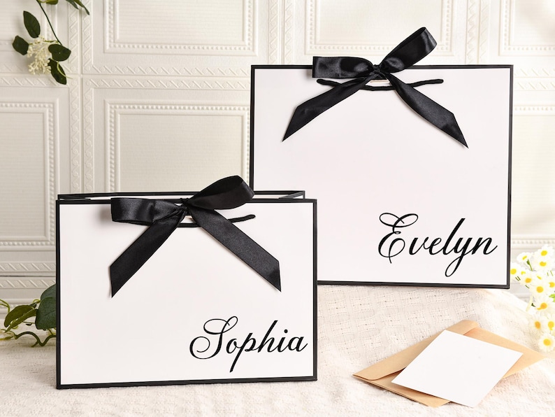 May include: Two white gift bags with black trim and black satin bows. One bag has the name "Sophia" in cursive, and the other has "Evelyn". The bags are on a white surface with a floral and card in the background.