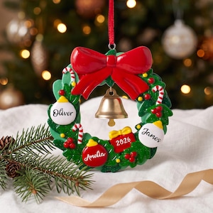 K&ouml;nnte beinhalten: Ein gr&uuml;ner Weihnachts-Kranz-Anh&auml;nger mit einer gro&szlig;en roten Schleife oben und einer goldenen Glocke in der Mitte. Der Kranz ist mit roten Beeren, Zuckerstangen und drei wei&szlig;en oder roten Ornamenten verziert, die mit den Namen Oliver, Amelia und James personalisiert sind, sowie einer roten Geschenkbox mit der Aufschrift 2026.