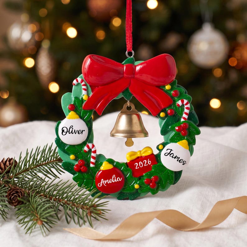 K&ouml;nnte beinhalten: Ein gr&uuml;ner Weihnachts-Kranz-Anh&auml;nger mit einer gro&szlig;en roten Schleife oben und einer goldenen Glocke in der Mitte. Der Kranz ist mit roten Beeren, Zuckerstangen und drei wei&szlig;en oder roten Ornamenten verziert, die mit den Namen Oliver, Amelia und James personalisiert sind, sowie einer roten Geschenkbox mit der Aufschrift 2026.