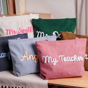 May include: A collection of six corduroy tote bags in various colors including cream, dark green, dark gray, light gray, pink, and brown. Each bag features a large, raised, white or light-colored script name or title, such as Mr. Smith, My Teacher, and others.