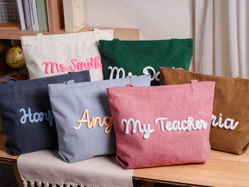 May include: A collection of six corduroy tote bags in various colors including cream, dark green, dark gray, light gray, pink, and brown. Each bag features a large, raised, white or light-colored script name or title, such as Mr. Smith, My Teacher, and others.
