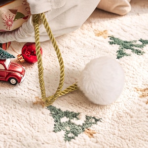 May include: A white fabric drawstring bag featuring a gold twisted rope cord and a large white faux fur pom-pom. The bag is decorated with a pattern of small green trees and is accompanied by a red toy car and a red Christmas ornament.