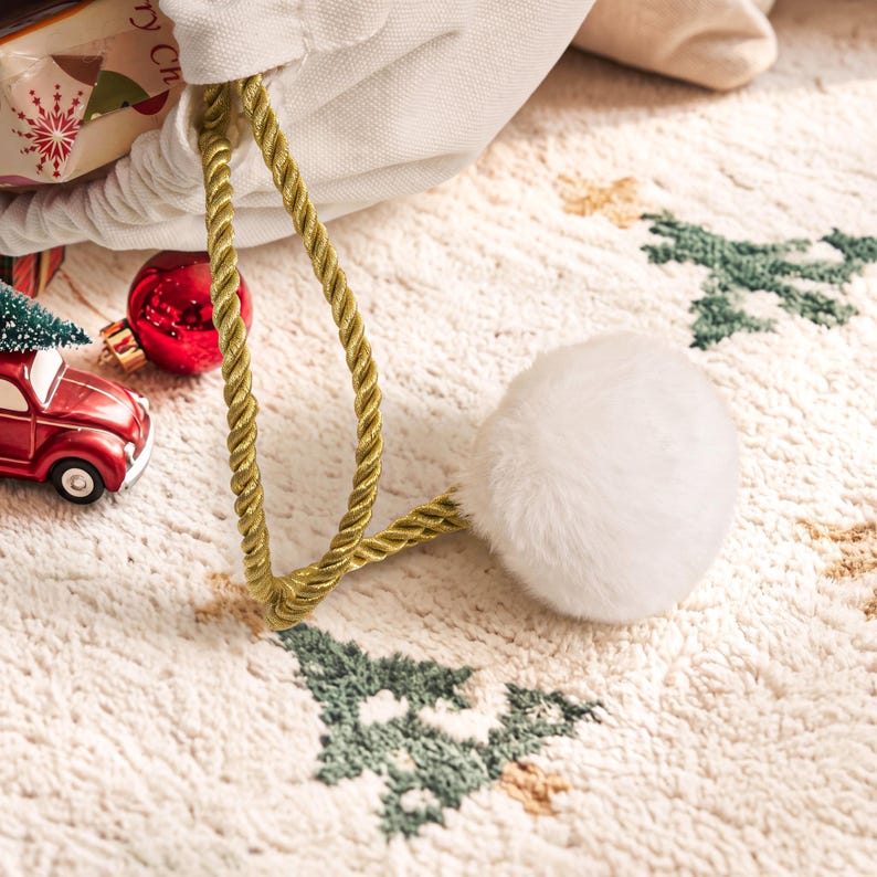 May include: A white fabric drawstring bag featuring a gold twisted rope cord and a large white faux fur pom-pom. The bag is decorated with a pattern of small green trees and is accompanied by a red toy car and a red Christmas ornament.
