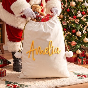 May include: A large off-white fabric gift sack featuring the name Amelia in yellow cursive lettering. The bag has gold drawstring cords with white pom-poms at the ends. A brown teddy bear and a small red toy truck are visible inside the opening.