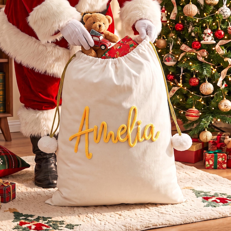 May include: A large off-white fabric gift sack featuring the name Amelia in yellow cursive lettering. The bag has gold drawstring cords with white pom-poms at the ends. A brown teddy bear and a small red toy truck are visible inside the opening.