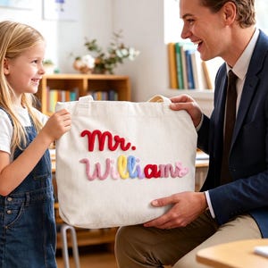 May include: A white corduroy tote bag featuring the text Mr. Williams embroidered in colorful, raised yarn lettering. The word Mr. is in red, while the name Williams is spelled out in alternating pink, yellow, blue, and red yarn letters.