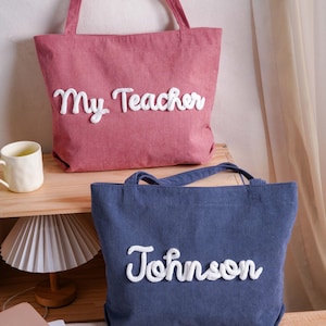 May include: A pink corduroy tote bag with the white embroidered text My Teacher and a blue corduroy tote bag with the white embroidered text Johnson. Both bags feature two shoulder straps and a textured fabric surface with vertical ribbing.