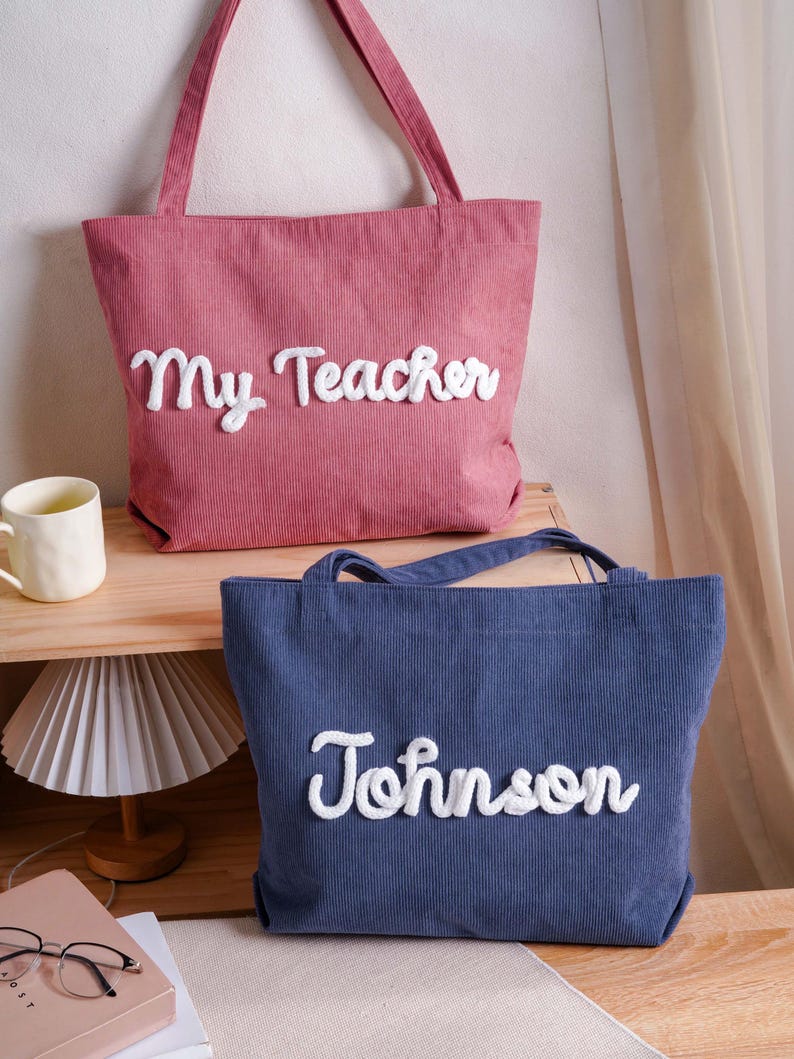 May include: A pink corduroy tote bag with the white embroidered text My Teacher and a blue corduroy tote bag with the white embroidered text Johnson. Both bags feature two shoulder straps and a textured fabric surface with vertical ribbing.