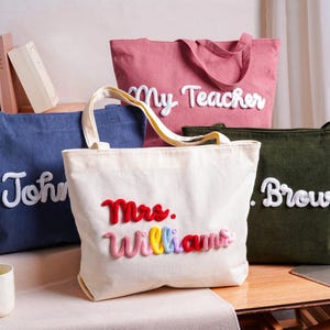 May include: Four corduroy tote bags in blue, pink, cream, and olive green. Each bag features raised, white or multicolored yarn-stitched lettering. The cream bag displays "Mrs. Williams" in red, pink, yellow, blue, and red yarn, while others show names.