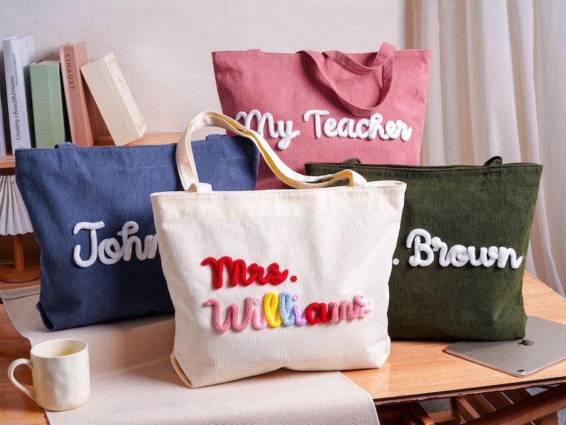 May include: Four corduroy tote bags in blue, pink, cream, and olive green. Each bag features raised, white or multicolored yarn-stitched lettering. The cream bag displays "Mrs. Williams" in red, pink, yellow, blue, and red yarn, while others show names.