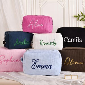 Op de afbeelding: Een verzameling gepersonaliseerde make-up tassen in verschillende kleuren, waaronder roze, marineblauw, wit, zwart, lichtblauw en bruin. Elke tas heeft een naam geborduurd in een contrasterende kleur, zoals "Alice", "Amelia" en "Camila".