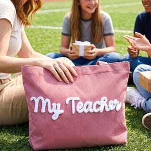 May include: A dusty rose corduroy tote bag featuring the white embroidered text My Teacher in a cursive font. The bag has two fabric handles and a textured ribbed surface, providing a spacious interior for carrying various items and essentials.