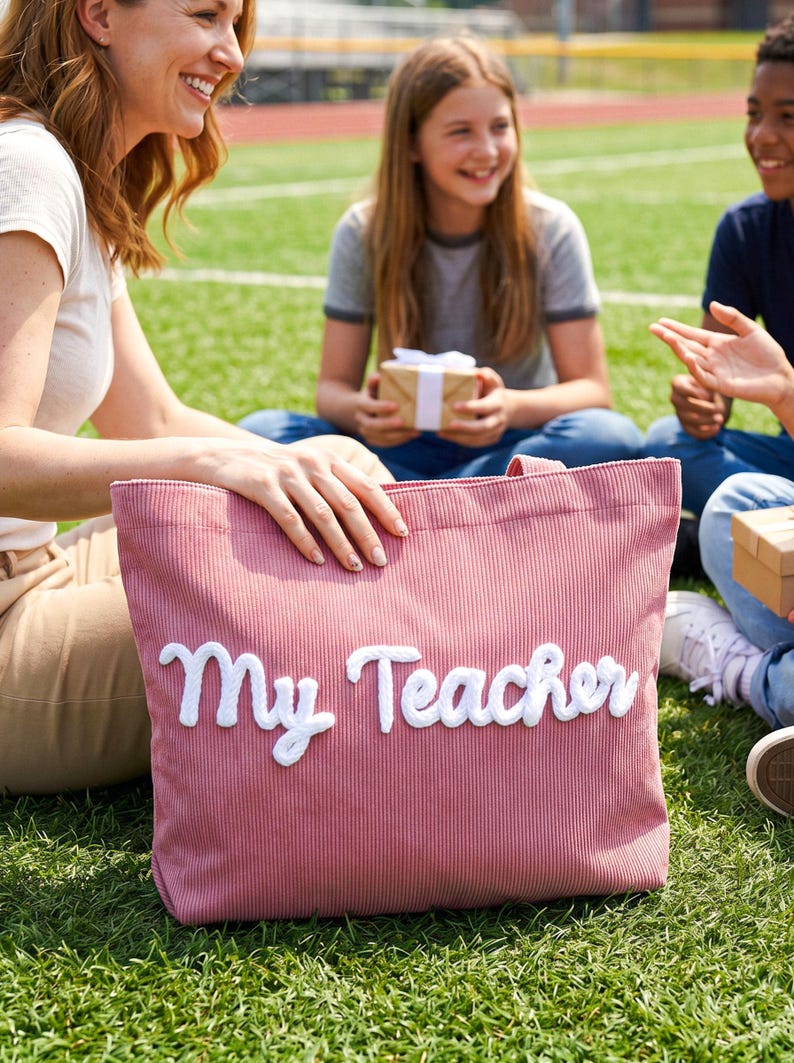 May include: A dusty rose corduroy tote bag featuring the white embroidered text My Teacher in a cursive font. The bag has two fabric handles and a textured ribbed surface, providing a spacious interior for carrying various items and essentials.