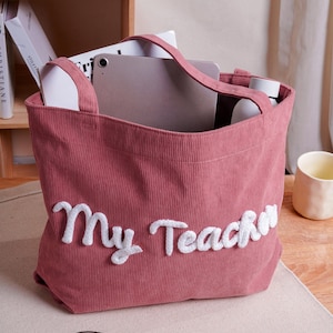 May include: A dusty rose corduroy tote bag featuring the white embroidered text "My Teacher" on the front. The bag has two matching fabric handles and is shown containing a tablet and a white bottle, with a pair of glasses resting in front.