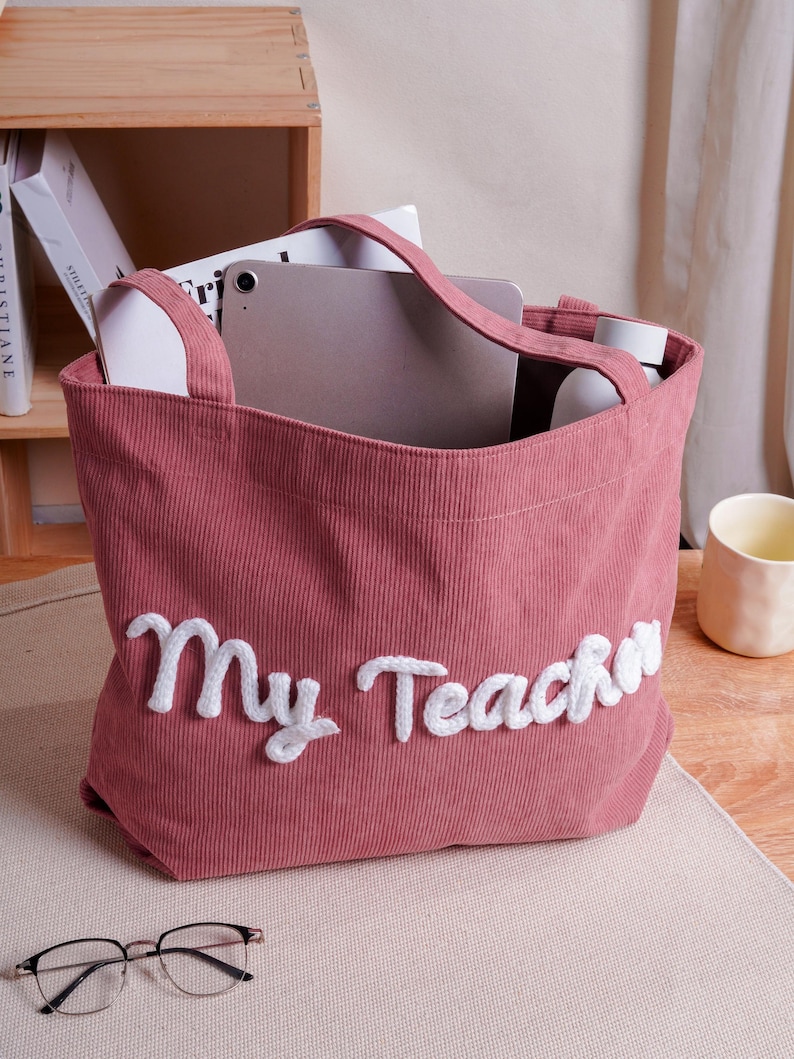 May include: A dusty rose corduroy tote bag featuring the white embroidered text "My Teacher" on the front. The bag has two matching fabric handles and is shown containing a tablet and a white bottle, with a pair of glasses resting in front.